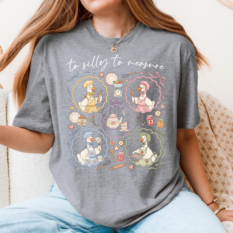 May include: A gray t-shirt featuring a whimsical design with the text "to silly to measure." The graphic showcases cartoon geese in chef hats and aprons, surrounded by kitchen tools and food items. The design is framed by colorful floral borders.