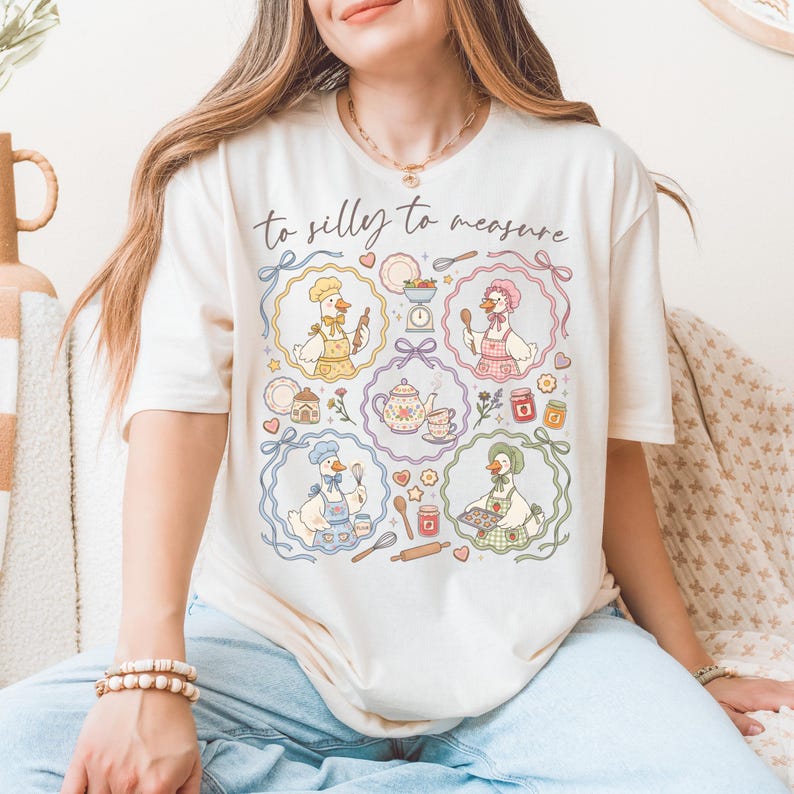 May include: Cream-colored t-shirt featuring a whimsical design with the text "to silly to measure." The graphic includes cartoon geese in chef hats and aprons, surrounded by baking-themed illustrations like teacups, rolling pins, and jars of jam.