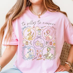 May include: Pink t-shirt with a whimsical design featuring cartoon geese in chef hats and aprons, surrounded by kitchen tools and food items. The text "to silly to measure" is written across the top.