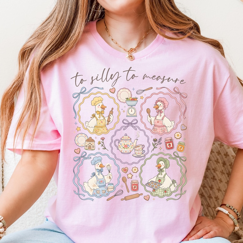 May include: Pink t-shirt with a whimsical design featuring cartoon geese in chef hats and aprons, surrounded by kitchen tools and food items. The text "to silly to measure" is written across the top.