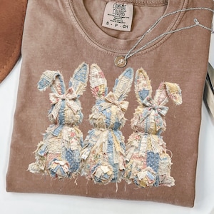 May include: A brown Comfort Colors t-shirt with three patchwork bunny rabbits. The bunnies are decorated with floral and lace patterns in blue, pink, and cream. A silver necklace with a small pendant is also visible.