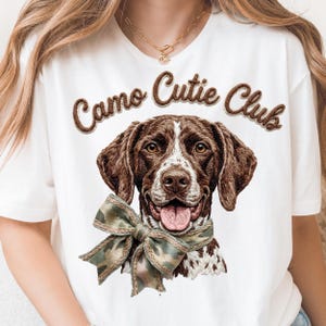 May include: White t-shirt featuring a detailed illustration of a brown and white dog wearing a camouflage bow. The text "Camo Cutie Club" is arched above the dog's head in brown lettering.