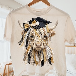 Könnte beinhalten: Cremefarbenes T-Shirt mit einer detaillierten Grafik einer Highland-Kuh, die einen Abschluss-Hut und eine dekorative Schleife trägt. Das Fell der Kuh ist mit Bändern in Schwarz, Gold und Creme verziert. Das Design beinhaltet eine goldene Quaste auf der Kappe.