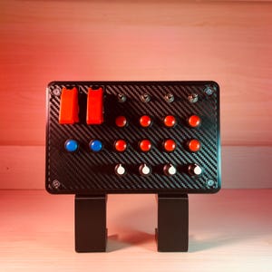 May include: A black panel with red, blue, and white buttons and toggle switches. The panel is mounted on a black stand with two legs.