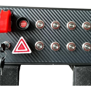 May include: A black control panel with a carbon fiber pattern. It features a red toggle switch, an "ENGINE START" button, a hazard light button, and several silver buttons. A key is also present.