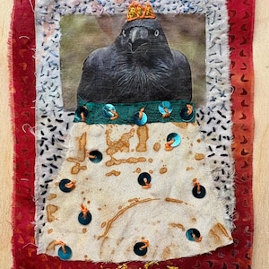 May include: Textile artwork depicting a crow wearing a crown and a green sequined collar. The crow is set against a fabric background with a red border. The text "TRUST THE PROCESS" is embroidered in gold thread.