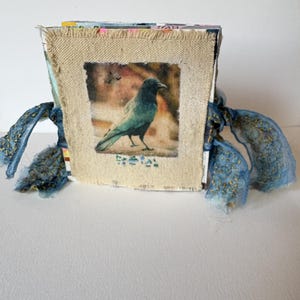 May include: A square book with a beige fabric cover featuring a printed image of a black crow. The cover has frayed edges and is tied on both sides with blue fabric ribbons that have gold-colored stitching details along the edges of the fabric.