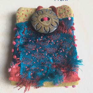 May include: A handmade textile art piece with a large wooden button. The piece features a combination of teal, coral, and yellow fabrics with intricate stitching and textured details. The word "Front" is written in blue.