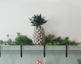 Puede incluir: Una repisa de chimenea blanca con una piña verde en la parte superior. Hay tres calcetines colgados de ganchos en la repisa. Los calcetines son blancos con detalles rojos y verdes. Hay una guirnalda verde con agujas de pino en la repisa.