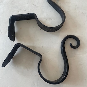 May include: Two black wrought iron curtain rod brackets. Each bracket has a curved, scroll-like design and a right-angled mounting plate with two holes for screws. The brackets are designed to hold a curtain rod.