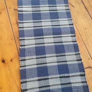 May include: A blue and gray woven runner rug with a checkered pattern. The rug features alternating bands of blue, gray, and black. The rug is rectangular and appears to be made of a durable material.