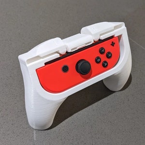 Könnte beinhalten: Ein weißes, 3D-gedrucktes Etui für einen Nintendo Switch-Controller. Das Etui hat zwei weiße Klappen, die sich herunterklappen lassen, um den Controller zu schützen. Der Controller ist rot mit schwarzen Knöpfen.