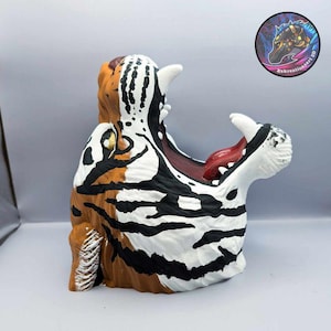 May include: A three-dimensional tiger head sculpture with its mouth wide open, featuring orange, black, and white fur patterns. The interior of the mouth is dark red with a visible tongue and white teeth. A circular logo with the text Kokreationsart 3D.