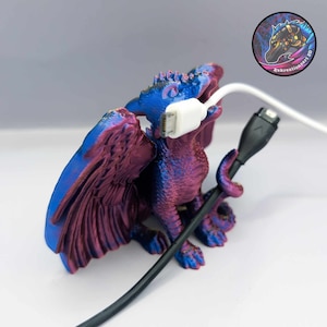 Puede incluir: Una figura de dragón impresa en 3D con un acabado iridiscente azul y morado, sosteniendo un cable de carga blanco en su boca. El dragón tiene alas y cola, y está sentado sobre una superficie blanca. Un cable de carga negro está envuelto alrededor de la cola del dragón.