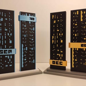 Könnte beinhalten: Vier abstrakte Skulpturen mit schwarzen rechteckigen Tafeln mit ausgeschnittenen Mustern. Zwei haben blaue Akzente mit den Buchstaben "SEP" und "18", die anderen beiden gelbe Akzente mit den Buchstaben "DEC" und "26". Sie stehen auf grauen und weißen Sockeln.