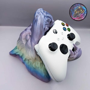 Może przedstawiać: Stojak na kontroler wydrukowany w 3D w kształcie tęczowego wilka z białym kontrolerem Xbox w środku. Pysk wilka jest otwarty, a kontroler jest trzymany w jego szczękach.