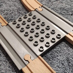 Könnte beinhalten: Ein graues Kunststoff-Adaptergleis mit einem zentralen Raster aus kreisförmigen Noppen zur Kompatibilität mit Bausteinen, verbunden mit zwei parallelen Holzeisenbahnschienen mit standardmäßigen männlichen und weiblichen Anschlüssen.