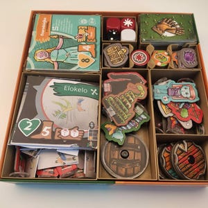 May include: A board game organizer with various game components. The box is divided into compartments holding cards, dice, and game pieces. The components include cards with illustrations, wooden tokens, and dice. The game appears to be fantasy-themed, with colorful artwork.