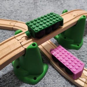 May include: A wooden train track set featuring a green plastic bridge support and a raised wooden track section. A green brick-compatible train car sits on the elevated track, while a pink brick-compatible train car rests on the lower track.