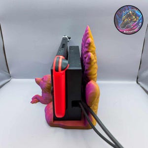 May include: A multi-colored 3D-printed Nintendo Switch dock featuring a fox design. The dock is painted in shades of pink, orange, and yellow. The Switch console and a red Joy-Con controller are inserted into the dock. The product is from Kekreationsart 3D.