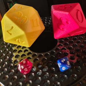May include: Three dice, one yellow, one red, and one blue, are sitting on a black metal grid. The yellow die has the numbers 1, 2, 4, 11, 12, and 20 on its faces. The red die has the numbers 3, 6, 8, and 9 on its faces. The blue die has the numbers 2, 6, and 7 on its faces.