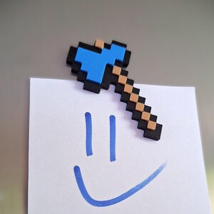 Könnte beinhalten: Ein pixeliger, axtförmiger Magnet mit einer leuchtend blauen Klinge, einem beigen Griff und einem schwarzen Umriss. Der Magnet befindet sich oben auf einem weißen quadratischen Blatt Papier, auf dem ein blaues, handgezeichnetes Smiley-Gesicht zu sehen ist.
