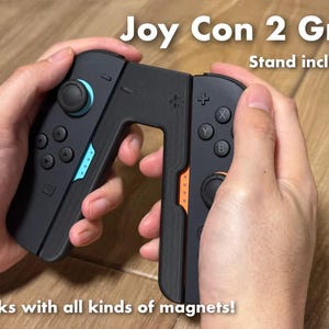 May include: Black Joy Con 2 Grip with blue and orange accents, held by hands. The text "Joy Con 2 Grip" and "Stand included" are visible. The text "Works with all kinds of magnets!" is also visible.
