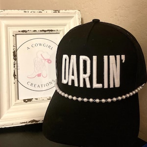 May include: Black trucker cap with the word "DARLIN'" embroidered in white. A string of white pearls is draped across the brim. A framed print with a cowgirl boot illustration is in the background.