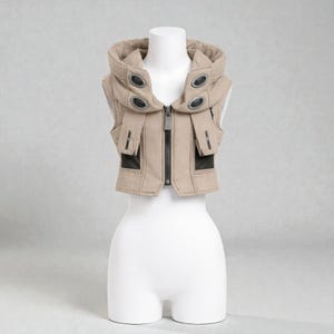 May include: A tan vest with a hood, displayed on a mannequin. The vest features a zip-up front, black accents, and decorative metal details. The hood has multiple oval-shaped metal accents. The vest is sleeveless and has a structured design.