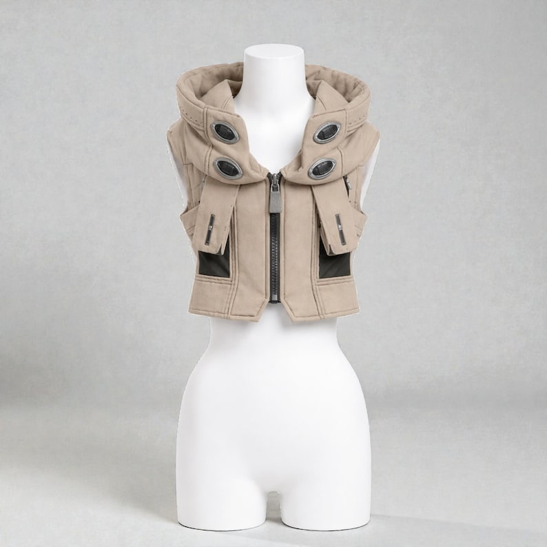 May include: A tan vest with a hood, displayed on a mannequin. The vest features a zip-up front, black accents, and decorative metal details. The hood has multiple oval-shaped metal accents. The vest is sleeveless and has a structured design.