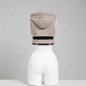 May include: A beige hooded vest with a black waistband and belt detail. The vest is sleeveless and has a zipper closure. The garment is displayed on a white mannequin against a neutral background.