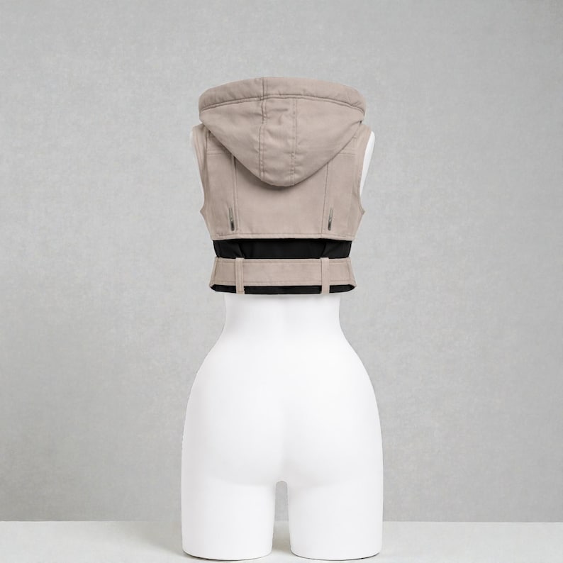 May include: A beige hooded vest with a black waistband and belt detail. The vest is sleeveless and has a zipper closure. The garment is displayed on a white mannequin against a neutral background.
