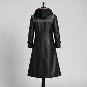 May include: A long, black leather coat with a hood. The coat has a tailored fit, buttoned cuffs, and a grey stripe across the upper back. The hood has an orange trim. The coat is displayed on a black mannequin.