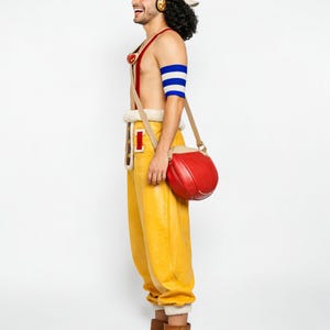 May include: A cosplay outfit featuring yellow pants with a white trim, a red and beige shoulder bag, and a red top with suspenders. The outfit includes a blue and white arm band. The person is wearing a headset and a wig.
