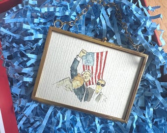 May include: A framed watercolor painting of three people, two men and one woman, with the American flag in the background. The men are wearing suits and the woman is wearing a dress. The painting is in a gold frame.