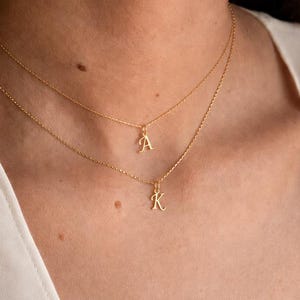 Pode incluir: Dois colares delicados em tom dourado com pequenos pingentes de letras pendentes. O colar superior exibe a letra A, enquanto o colar inferior apresenta a letra K. Ambas as correntes são finas e metálicas, repousando sobre o pescoço.