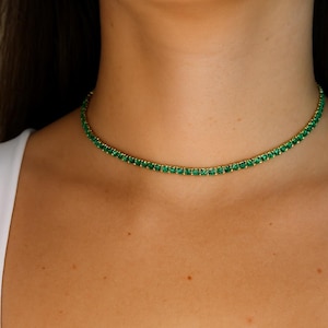Puede incluir: Un delicado collar gargantilla con una fila de pequeñas y vibrantes piedras preciosas verdes engastadas en metal dorado. El collar se asienta con gracia en el cuello, creando un accesorio sutil pero llamativo. El diseño es simple y elegante.