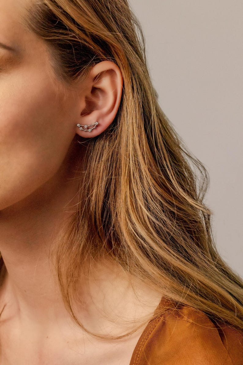 May include: A pair of silver earrings with a delicate chain design. The earrings are shaped like a crescent moon and feature small, sparkling stones.