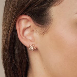 May include: A close-up of a gold-colored flower-shaped stud earring with pink and clear gemstones. The earring is worn on a person's ear. The earring has a delicate, feminine design.