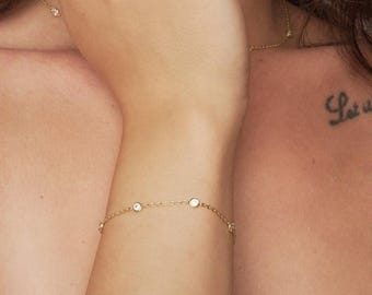 May include: Delicate gold bracelet featuring several clear, round gemstones. The bracelet is worn on the wrist and is connected by a fine gold chain. A matching necklace with similar gemstones is also visible.