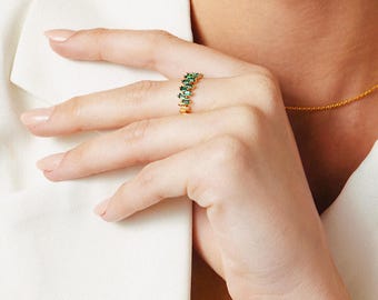 May include: A gold-colored ring with emerald-green rectangular stones set in a row. The ring is on a finger, and the hand is resting on a white surface. A gold necklace is also visible.