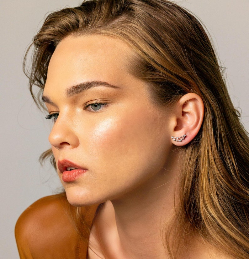 May include: A woman with long brown hair wearing a small silver earring with a delicate chain design.