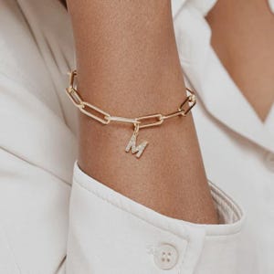 May include: Gold chain bracelet with a charm in the shape of the letter M, encrusted with small clear stones.
