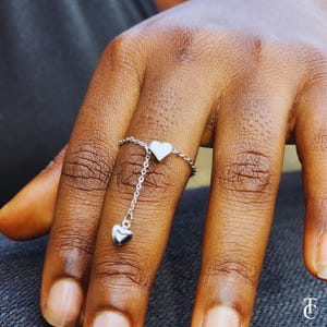 May include: A silver ring with a dangling heart charm. The ring is on a finger and the charm is hanging down.