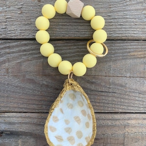 May include: A yellow beaded keyring with a gold key ring and a gold oyster shell charm. The oyster shell has a white interior with a brown speckled pattern.