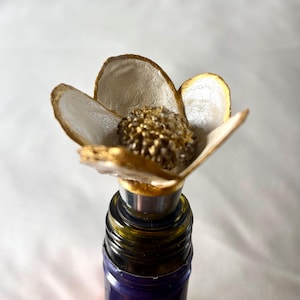 May include: A white and gold flower-shaped bottle stopper with a gold centre. The stopper is on top of a wine bottle.
