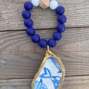 May include: A blue and white oyster shell charm with a gold rim and a blue and white fish design. The charm is attached to a blue silicone bracelet with white beads and a wooden bead.