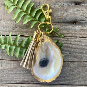 May include: Gold oyster shell keychain with a beige tassel. The oyster shell is painted gold and has a white interior. The keychain is attached to a gold ring and a gold lobster clasp.