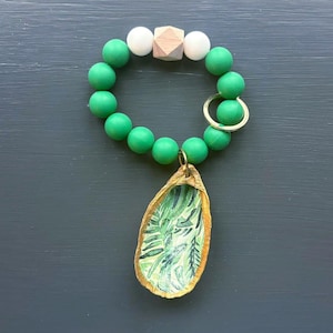 May include: A green beaded keyring with a wooden bead and white beads. The keyring has a gold ring and a gold charm with a green leaf design.