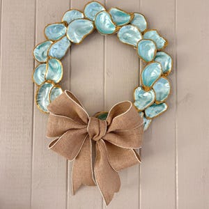 May include: A decorative wreath made of blue-green oyster shells with gold trim. A large burlap bow with white trim is attached to the bottom of the wreath. The wreath is displayed against a light gray wooden background.